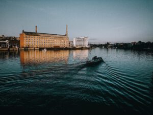 Im Abendlicht liegt die tiefblaue Spree auf der ein Boot fährt, von der untergehenden Sonne wird ein gelbes Industriegebäude angestahlt